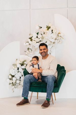 A touching moment captured in a bright studio with white flowers as two sit in an emerald green accent chair.
