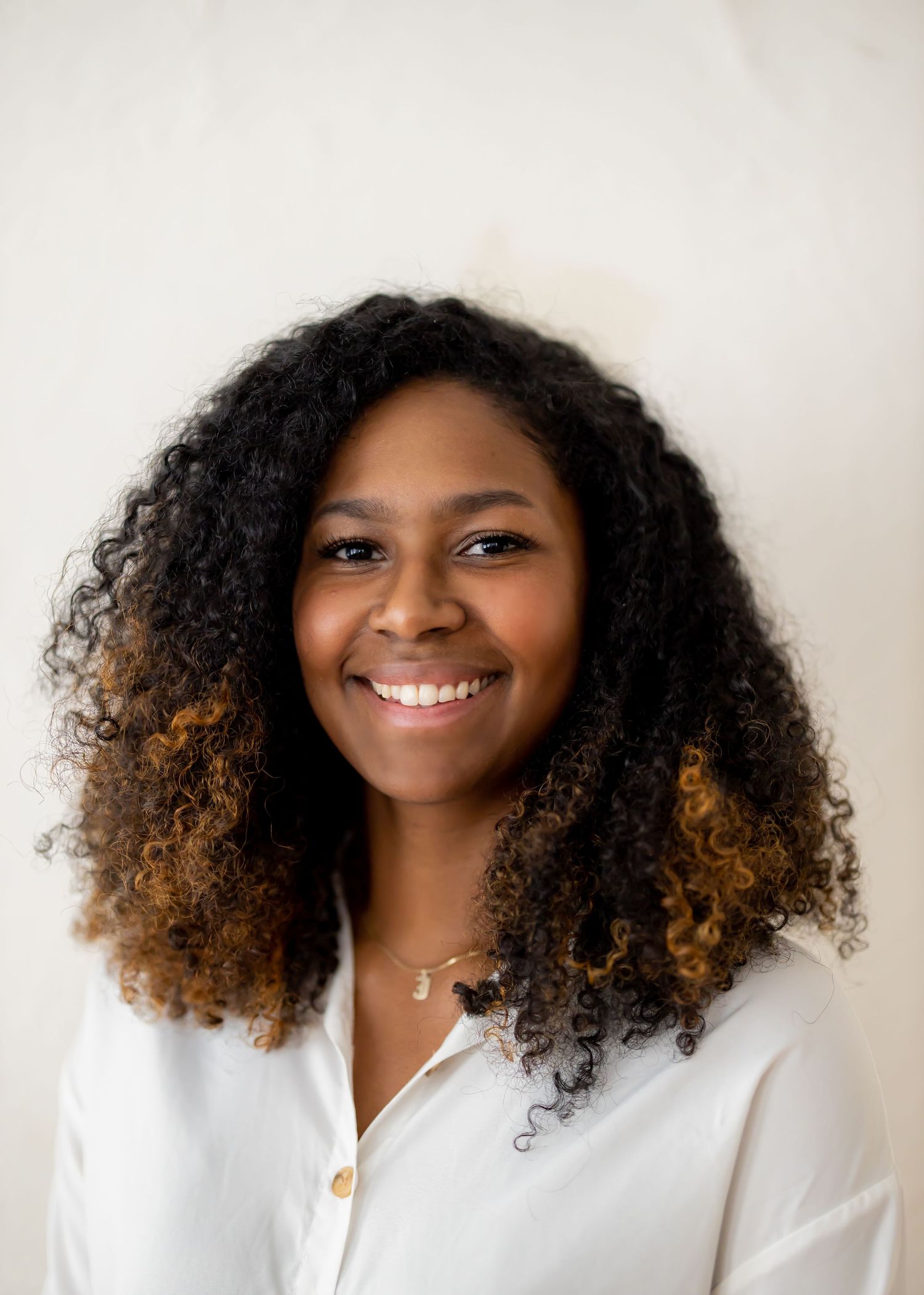 Professional headshot portrait showing a bright smile and voluminous curly hair woman with ombre coloring against a light background.