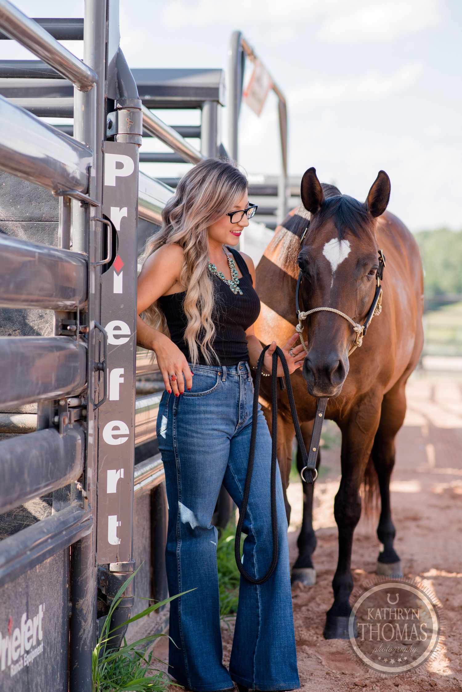 Someone in flared jeans and black top stands with a brown horse by metal stalls.