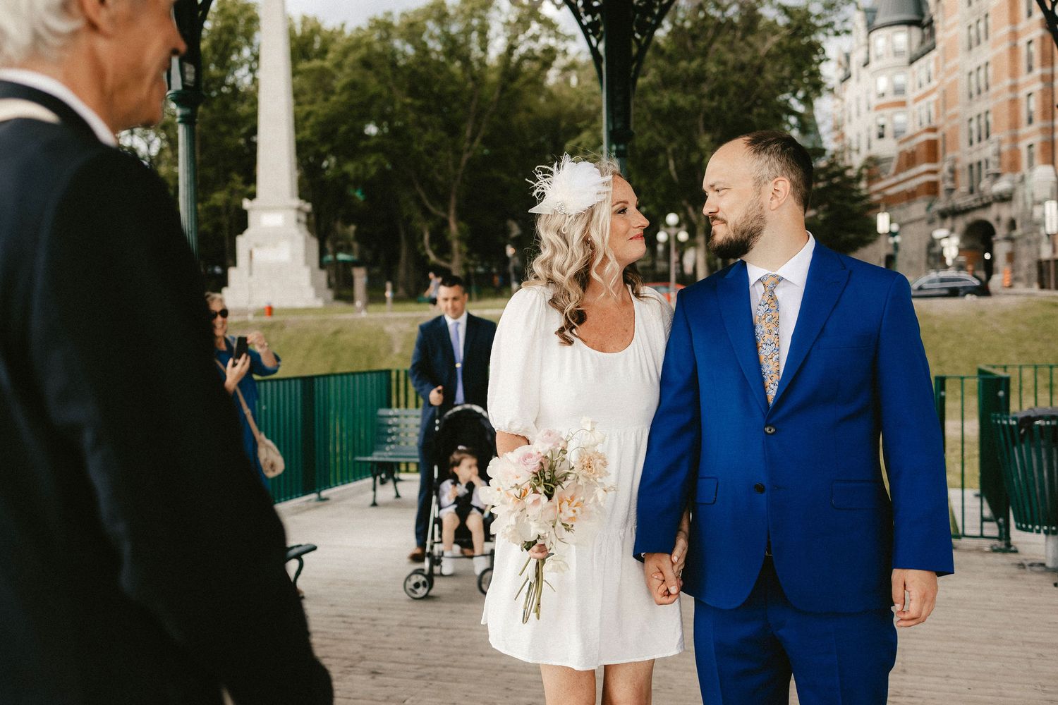 An outdoor wedding ceremony with a couple in white and blue attire near a monument.