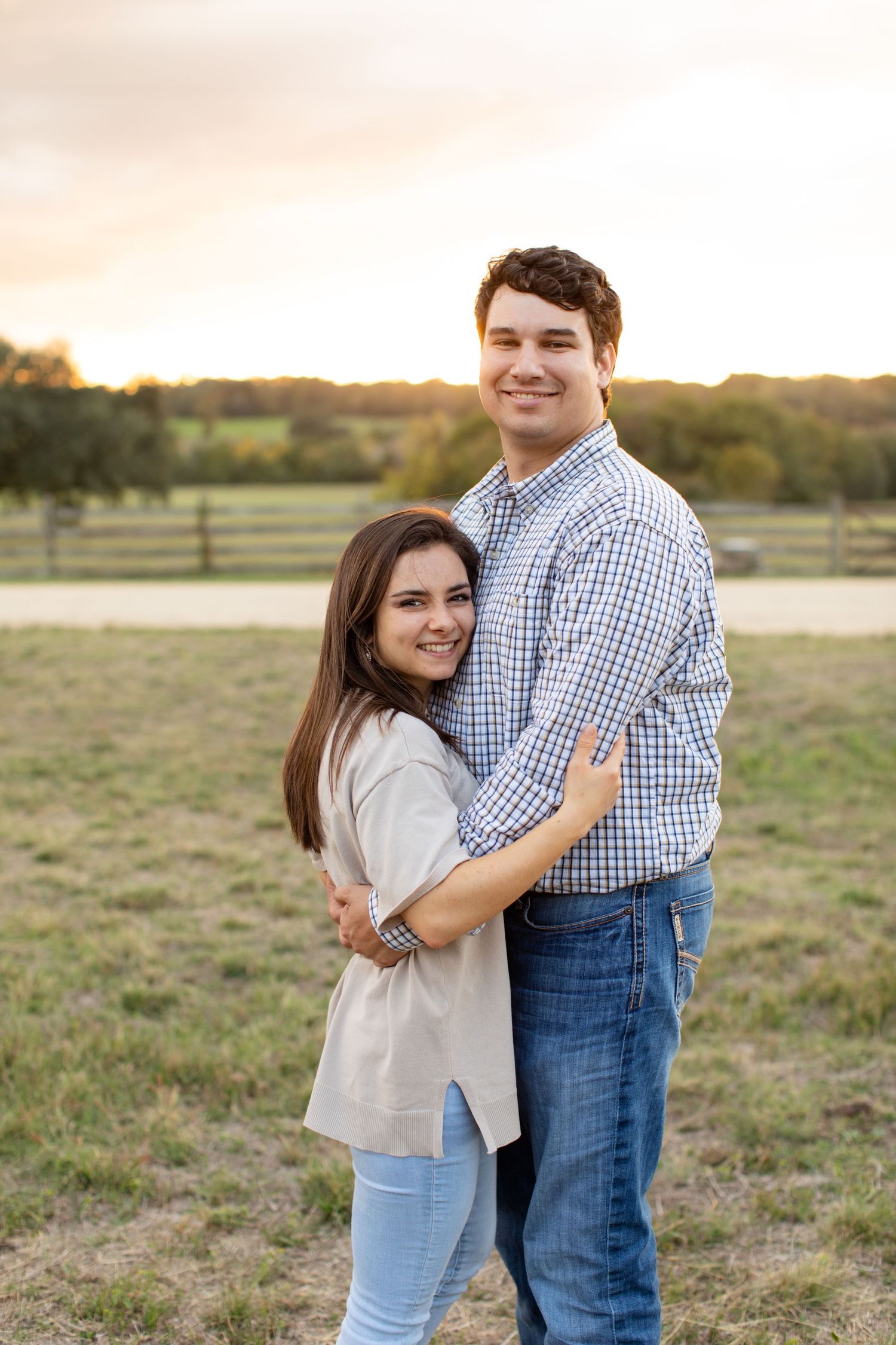Couple hugging while standing in a field at sunset with fenced pastures in the background.