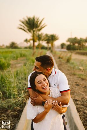 A couple laughing together on a path with palm trees and greenery in the background.
