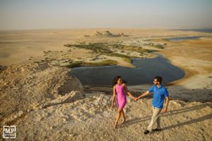 A couple holding hands while walking along a desert cliff overlooking a scenic oasis.