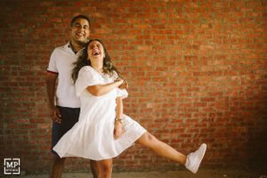 A playful couple poses against a brick wall, one lifting the other in a fun embrace.