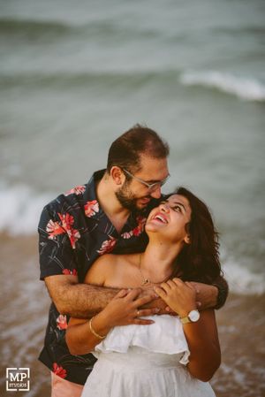 A joyful couple laughs together by the ocean, one wearing a floral shirt.