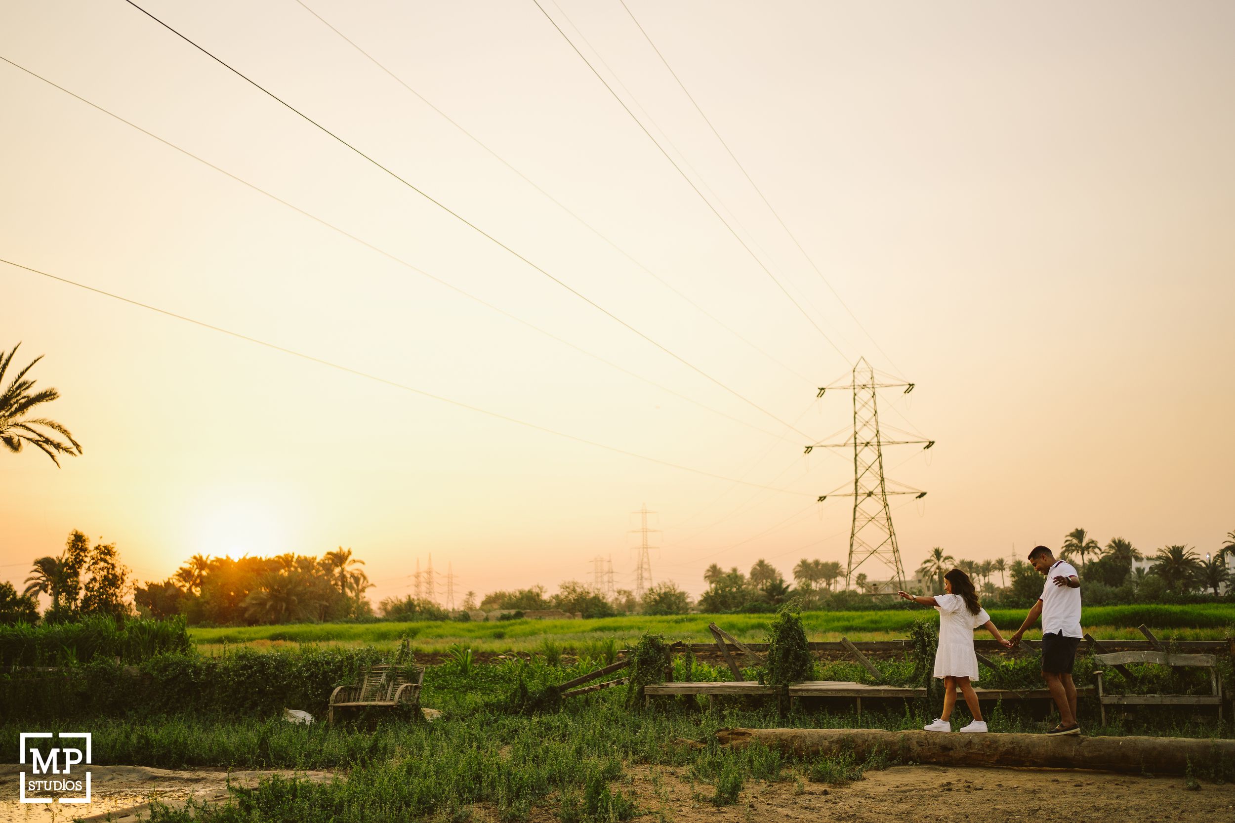 Two people stand in a field at sunset with power lines stretching across the golden sky.