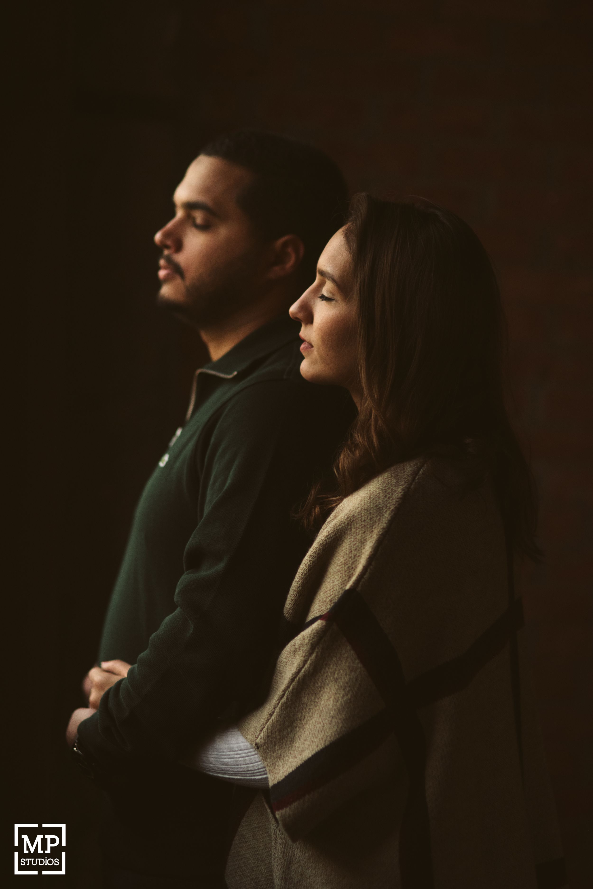 A couple in profile stands against a dark background in casual winter wear.