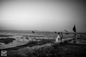 A couple stands on a rocky beach at sunset with flags waving in the distance.