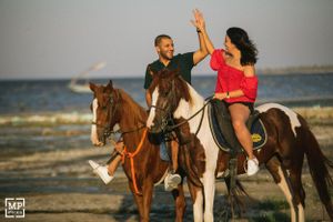 A couple high-fives while riding horses on a beach at sunset.