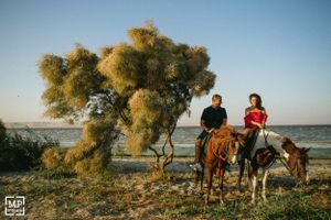 A couple rides horses beneath a large tree by the ocean.
