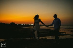A couple holds hands in silhouette against a dramatic orange sunset by the water.
