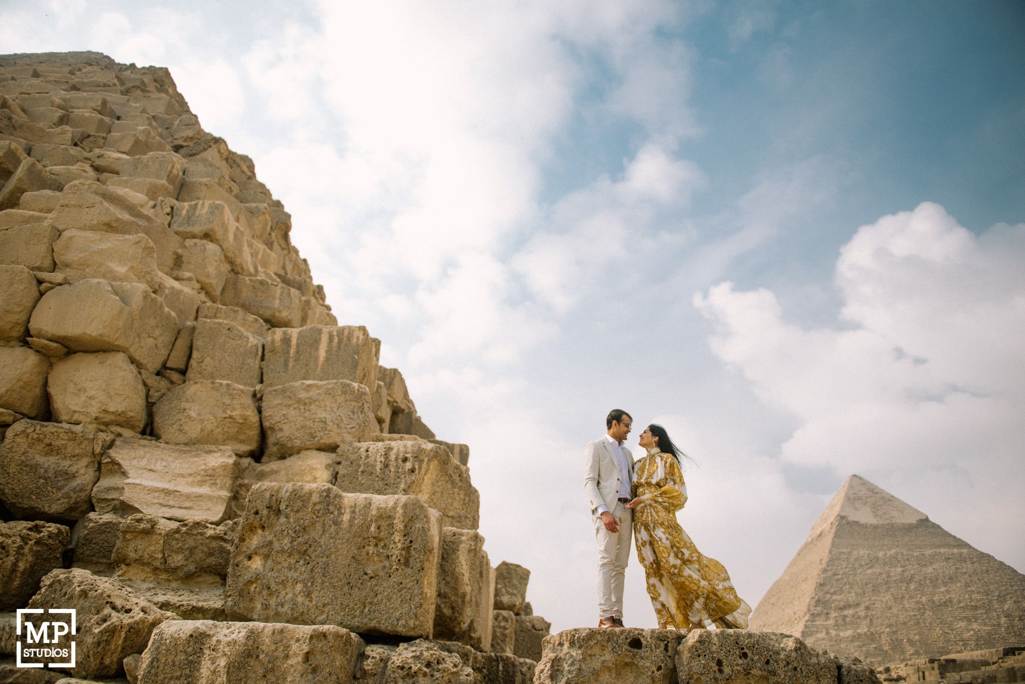 Romantic couple standing on ancient stone pyramid structure against cloudy sky.