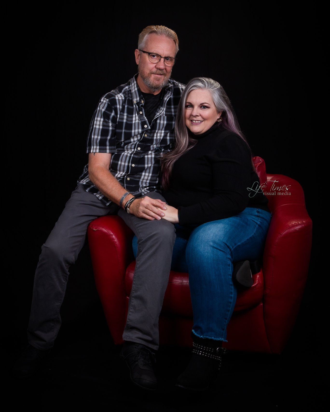 A couple sits together on a red leather chair against a black background in a studio portrait setting.