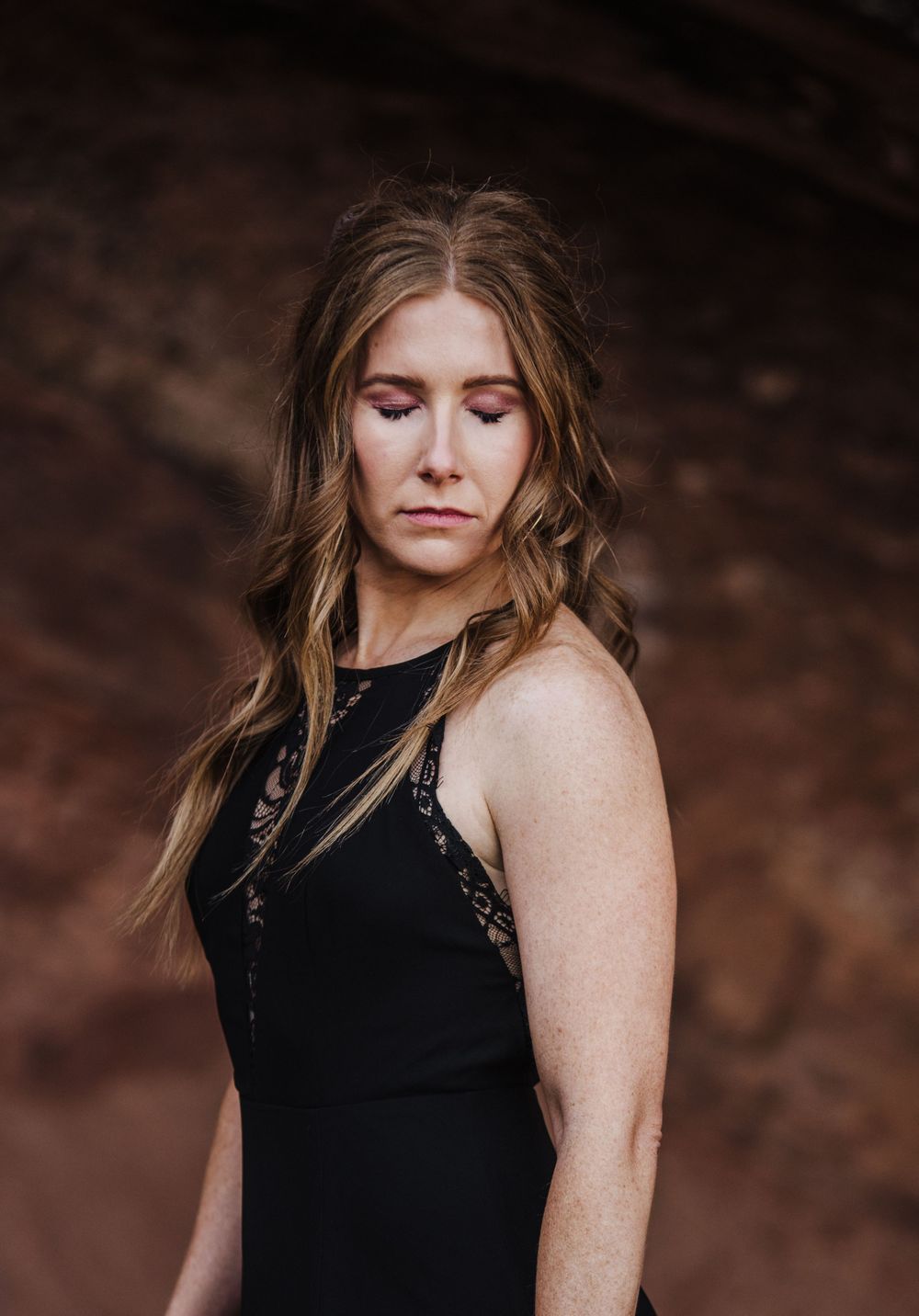 A figure in a sleeveless black dress stands against a dramatic red rock wall backdrop.