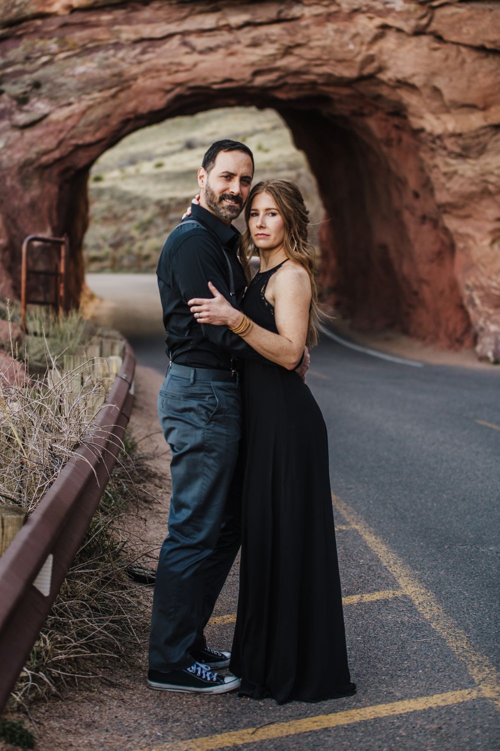 Romantic couple embrace in formal wear standing beneath a red rock tunnel.
