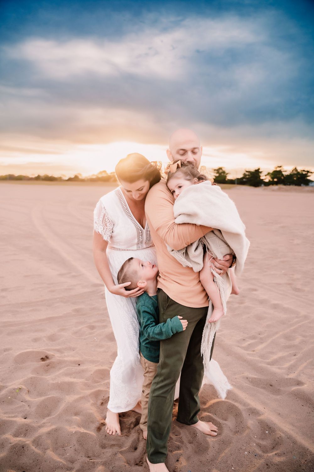 parents standing on south beach in Staten Island NY with dad holding young daughter and mom cuddling sons head