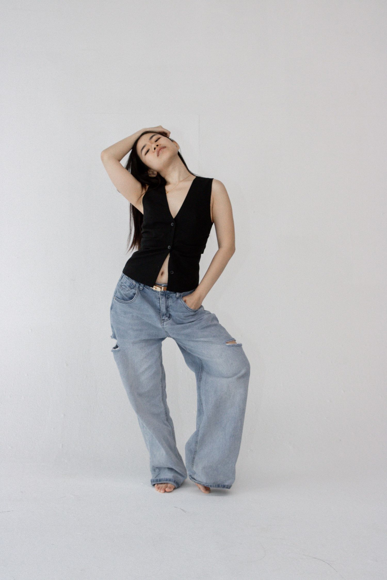 Casual fashion pose in black vest and wide-leg jeans against white studio backdrop.
