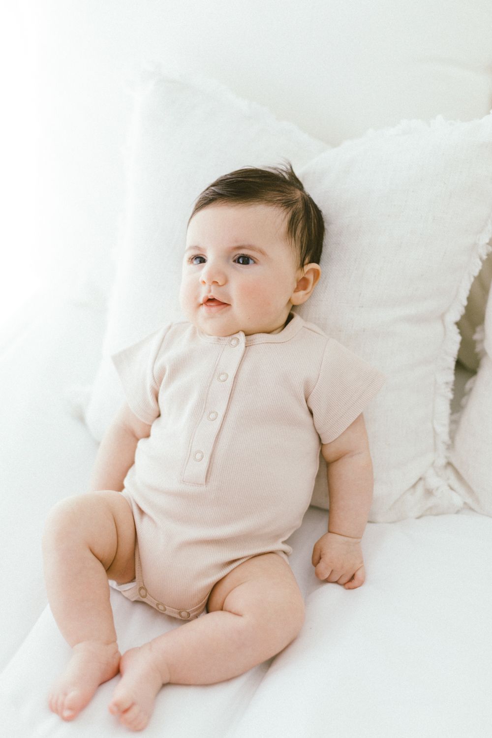 Baby wearing soft pink onesie lies contentedly on white bedding.