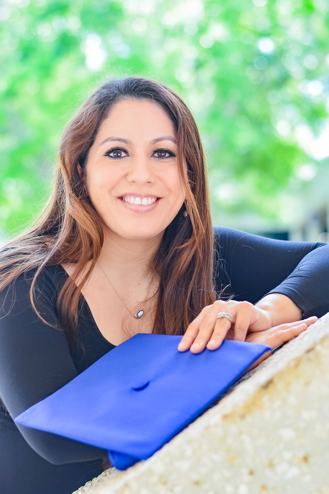 Graduate in cap and gown poses with blue diploma folder against blurred green background.