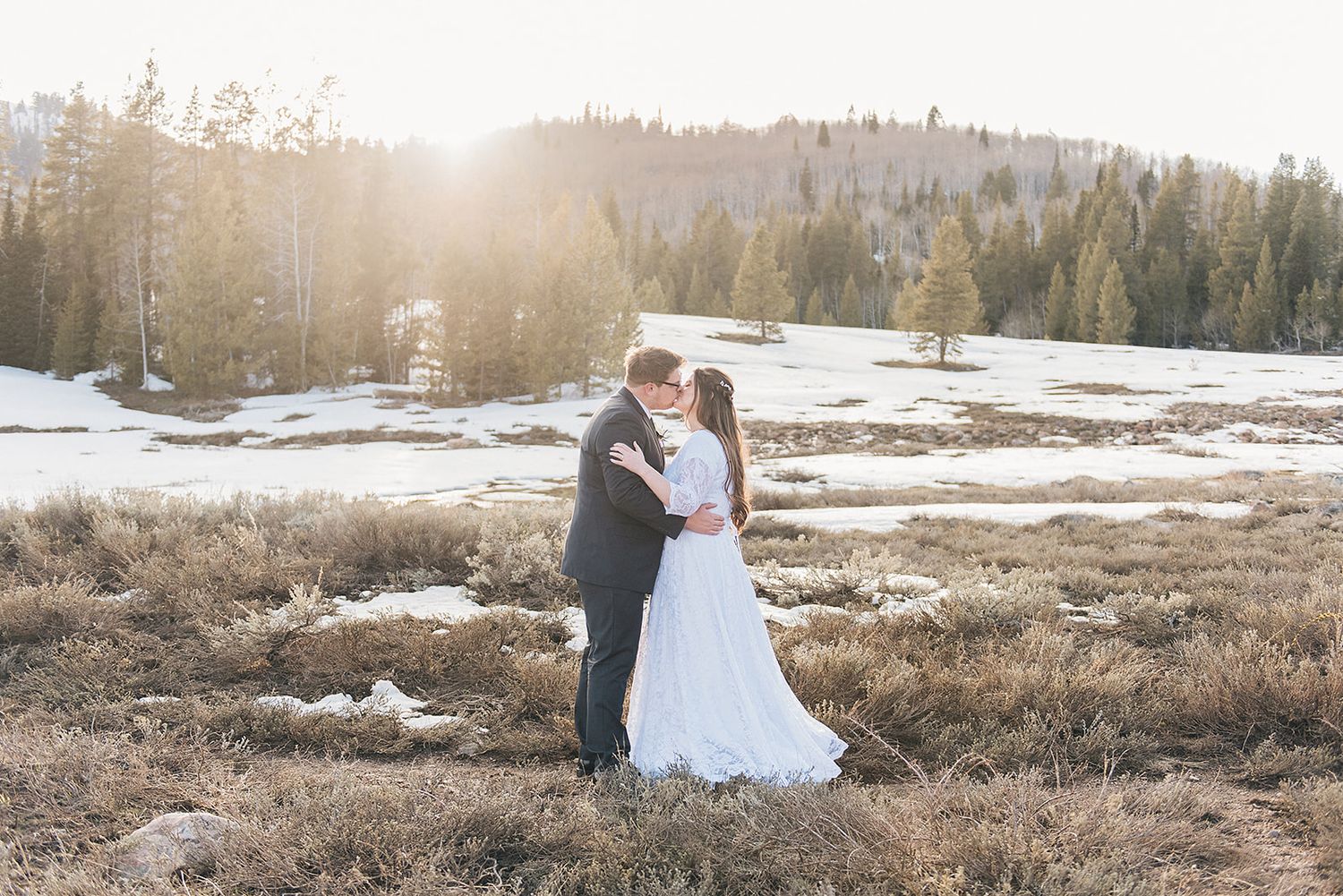 Sunlit wedding portrait in a mountain meadow with patches of snow and evergreen trees.