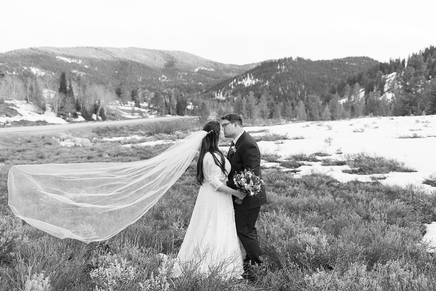 Black and white photo of newlyweds embracing in a winter mountain landscape with a flowing veil.