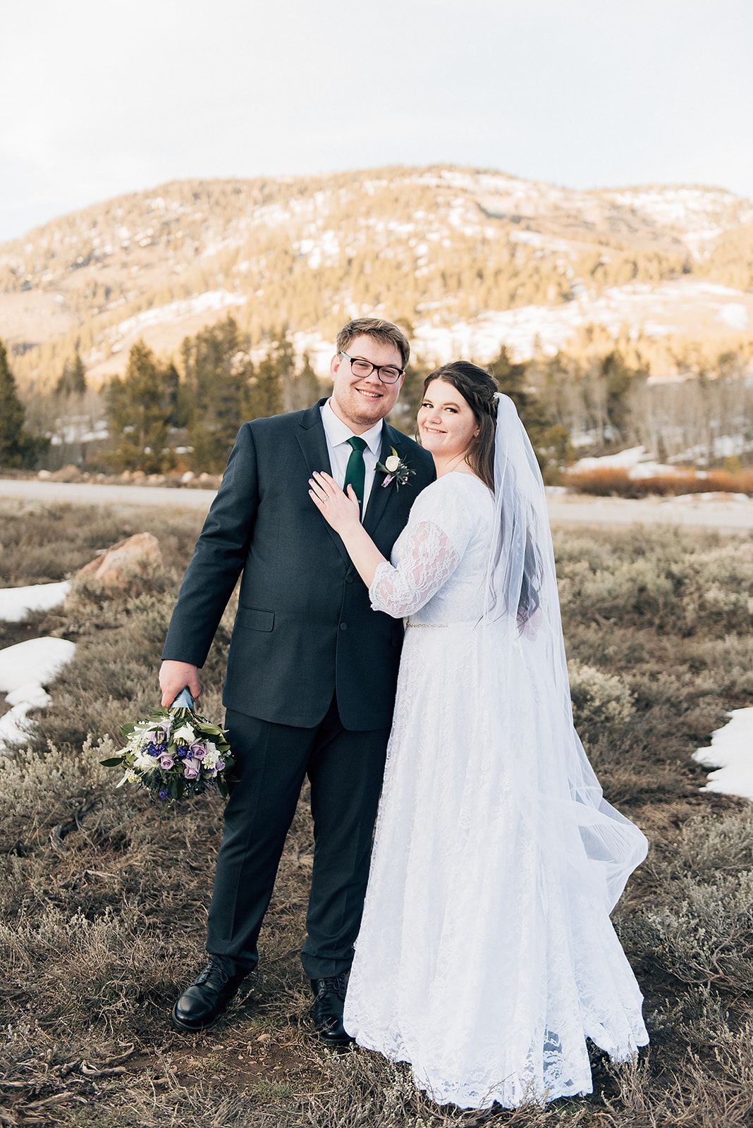 Newlyweds pose together holding a purple bouquet in a winter mountain setting with patches of snow.