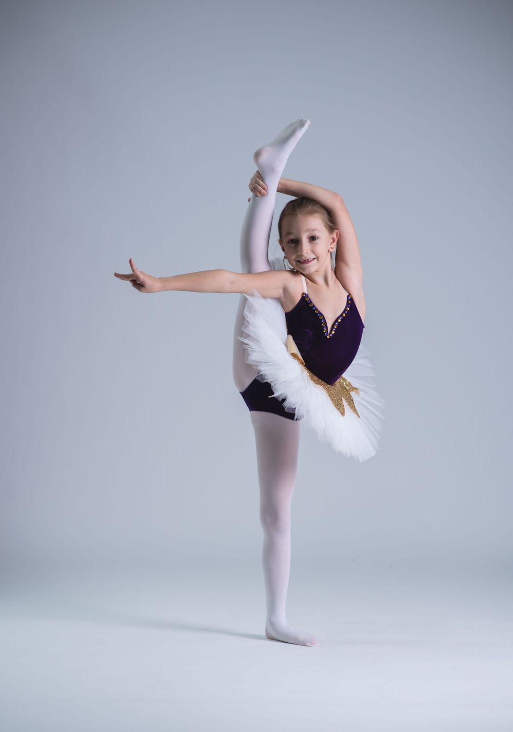 Young ballet dancer in purple leotard and white tutu performs graceful arabesque pose.