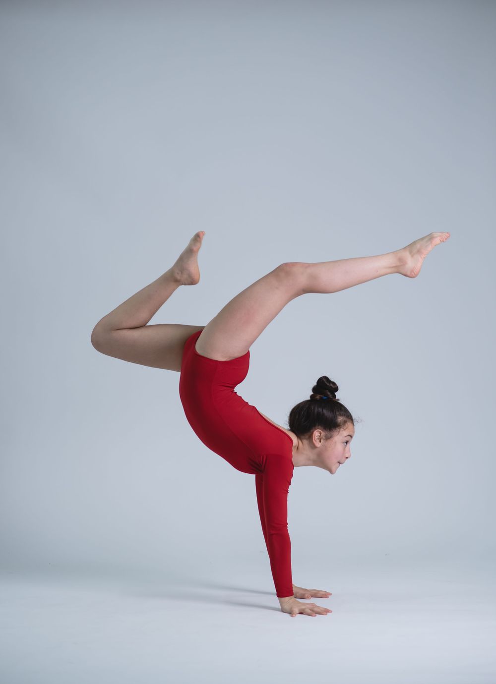 Gymnast in red leotard demonstrates impressive scorpion pose against grey background.