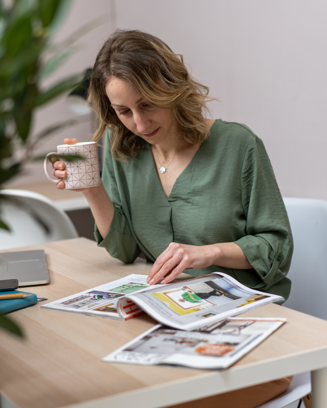 Woman in green blouse reading magazine while enjoying coffee at desk.