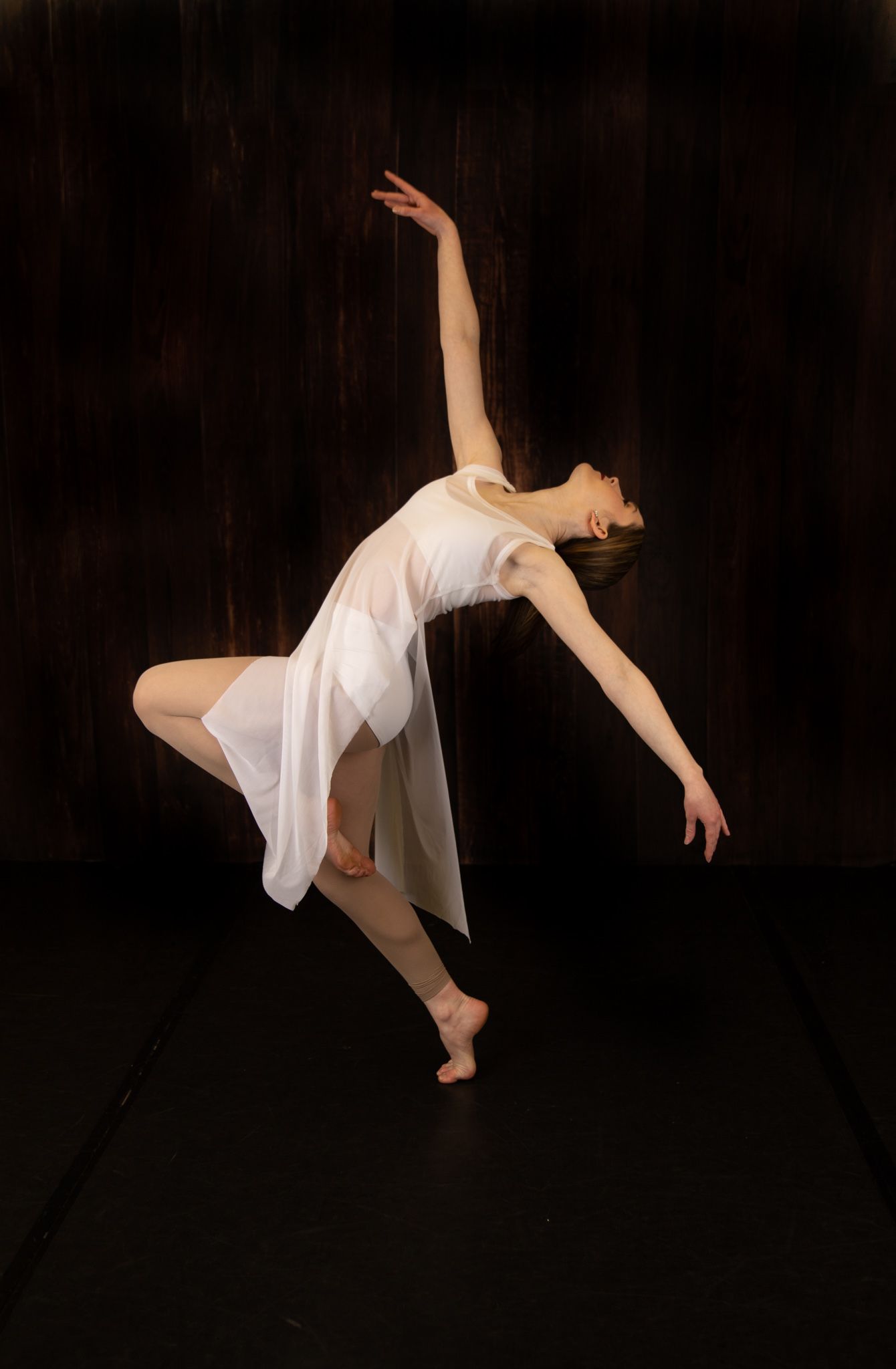 Dancer in white dress performs graceful movement against dark background.