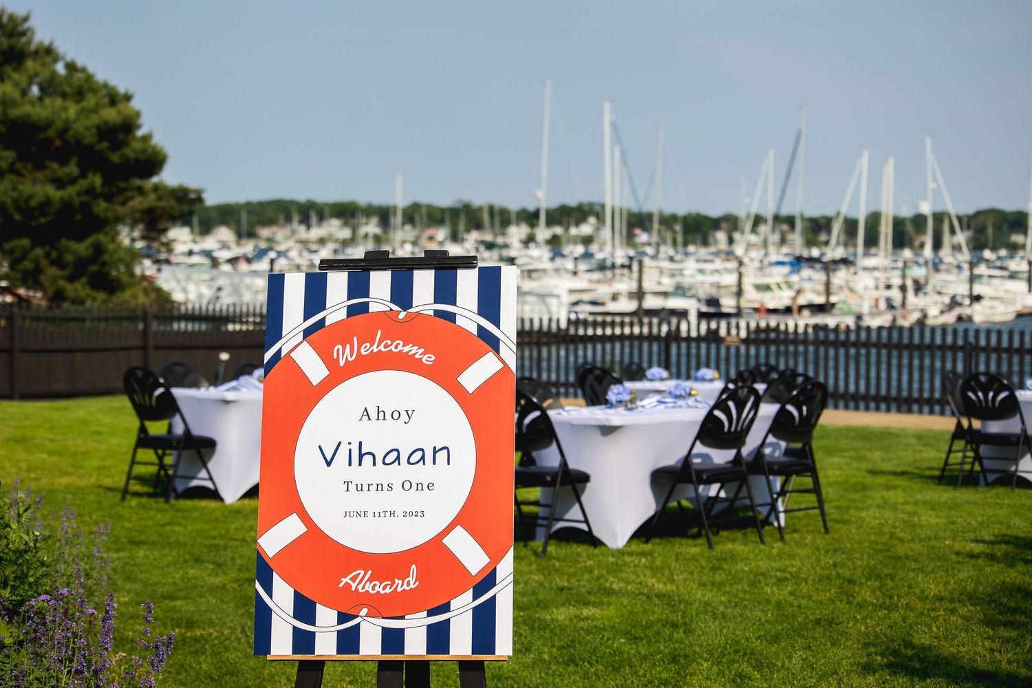 Outdoor event setup with striped sign and marina view featuring decorated tables and chairs on grass.