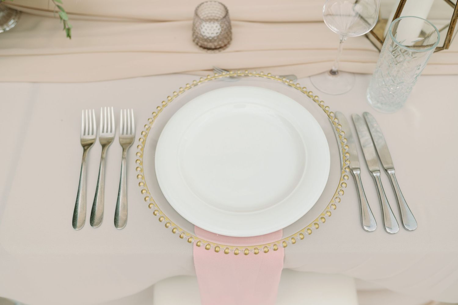 Place setting with white plate, beaded charger, and silver cutlery on blush tablecloth.