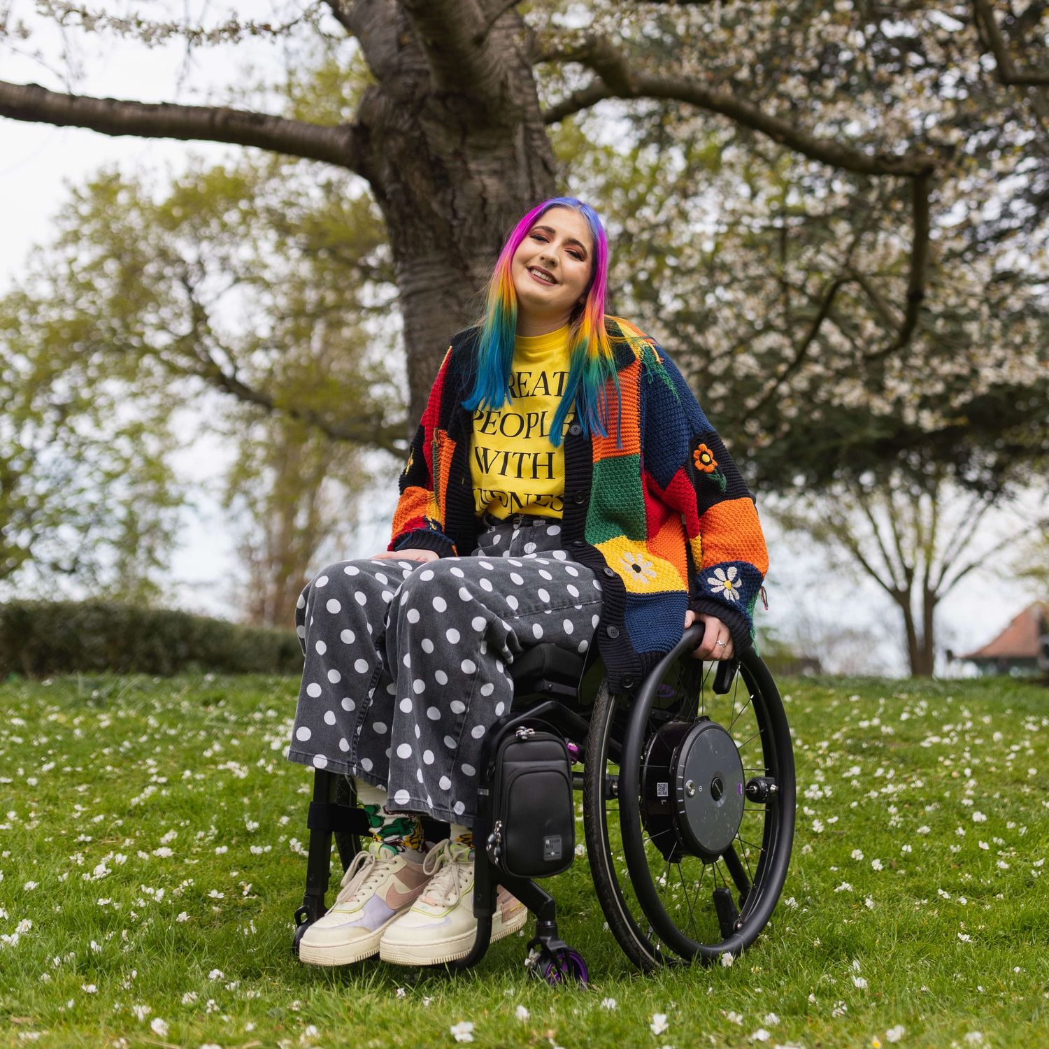London rainbow haired disabled influencer posing sat smiling in their wheelchair in a daisy field.