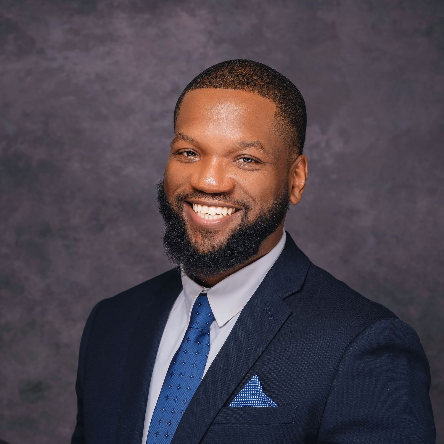Professional portrait of person in navy suit with blue tie and pocket square.