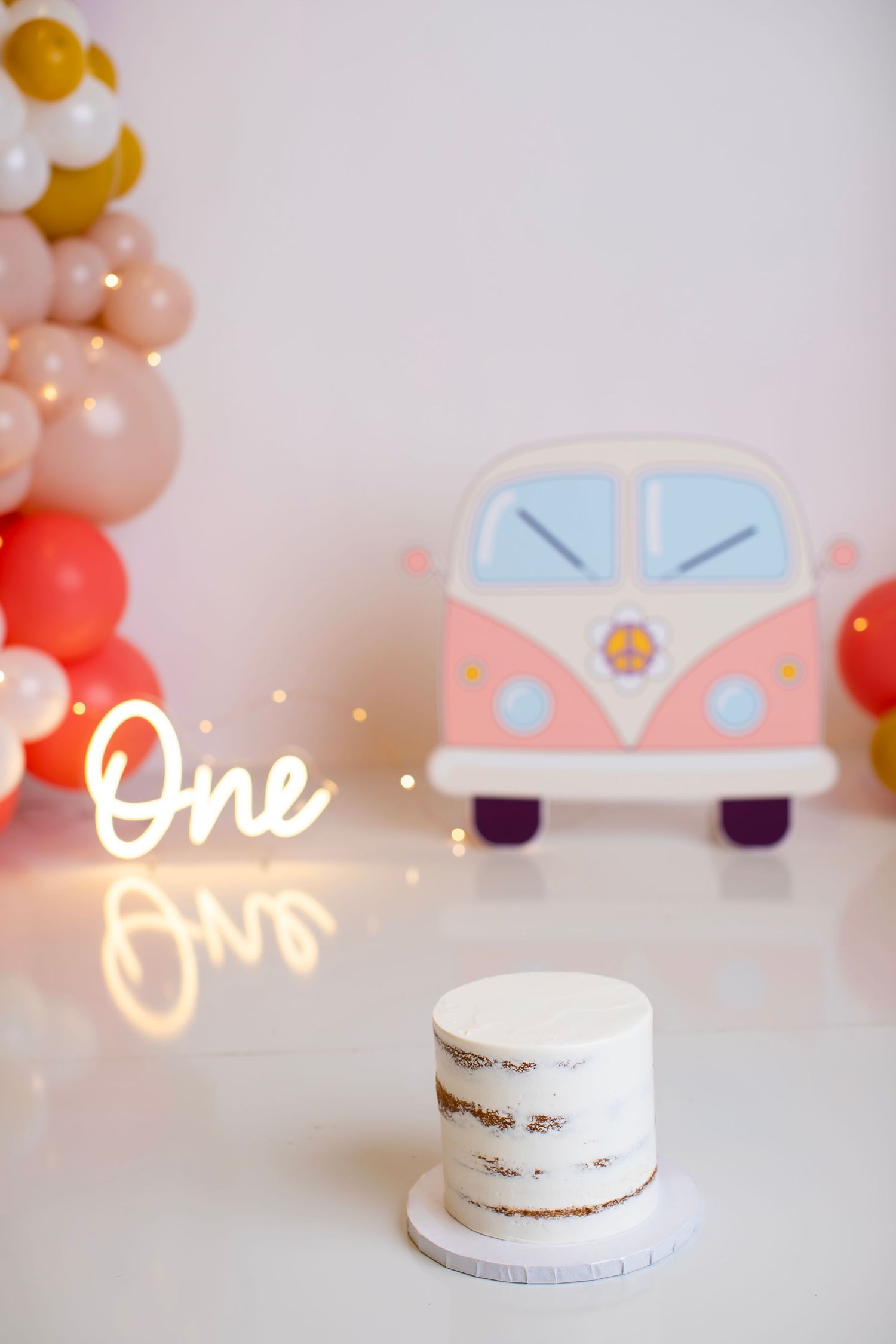 First birthday setup with white cake, neon 'One' sign, and retro van decoration against balloon arch.