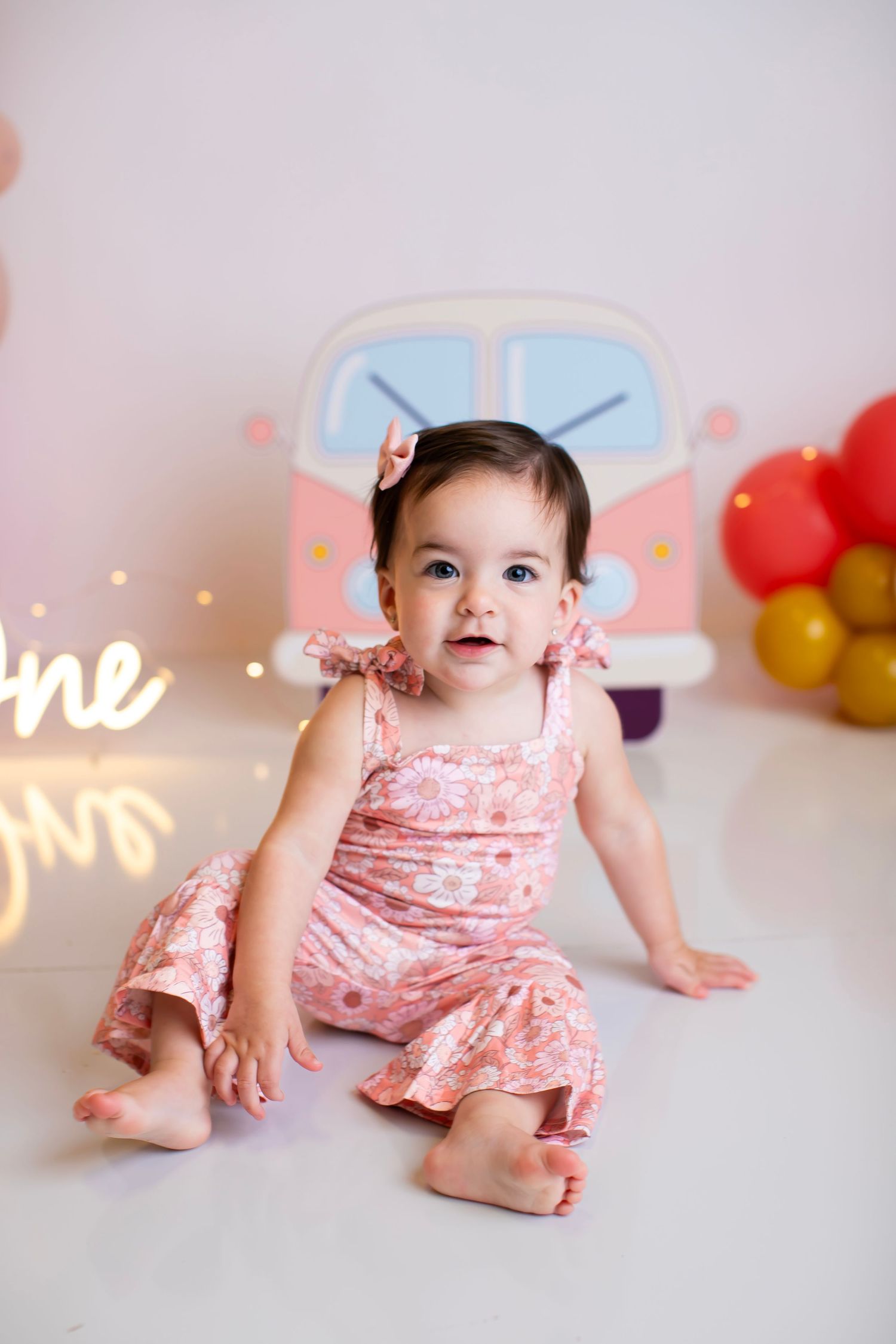 Baby in pink romper sitting on floor with vintage van decoration and balloon arch in background.
