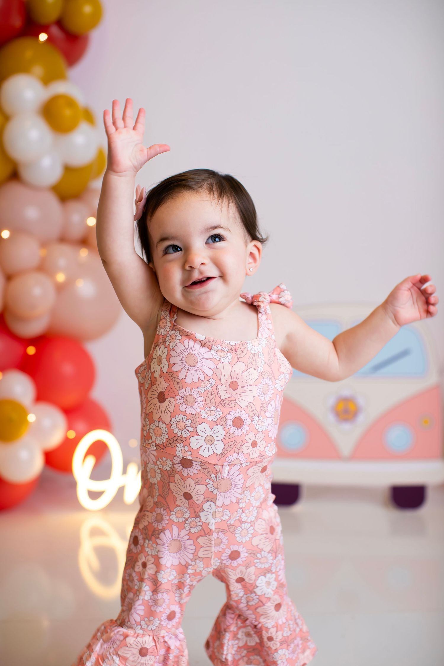 Baby in pink outfit raising hand with balloon arch and neon 'One' sign in background.