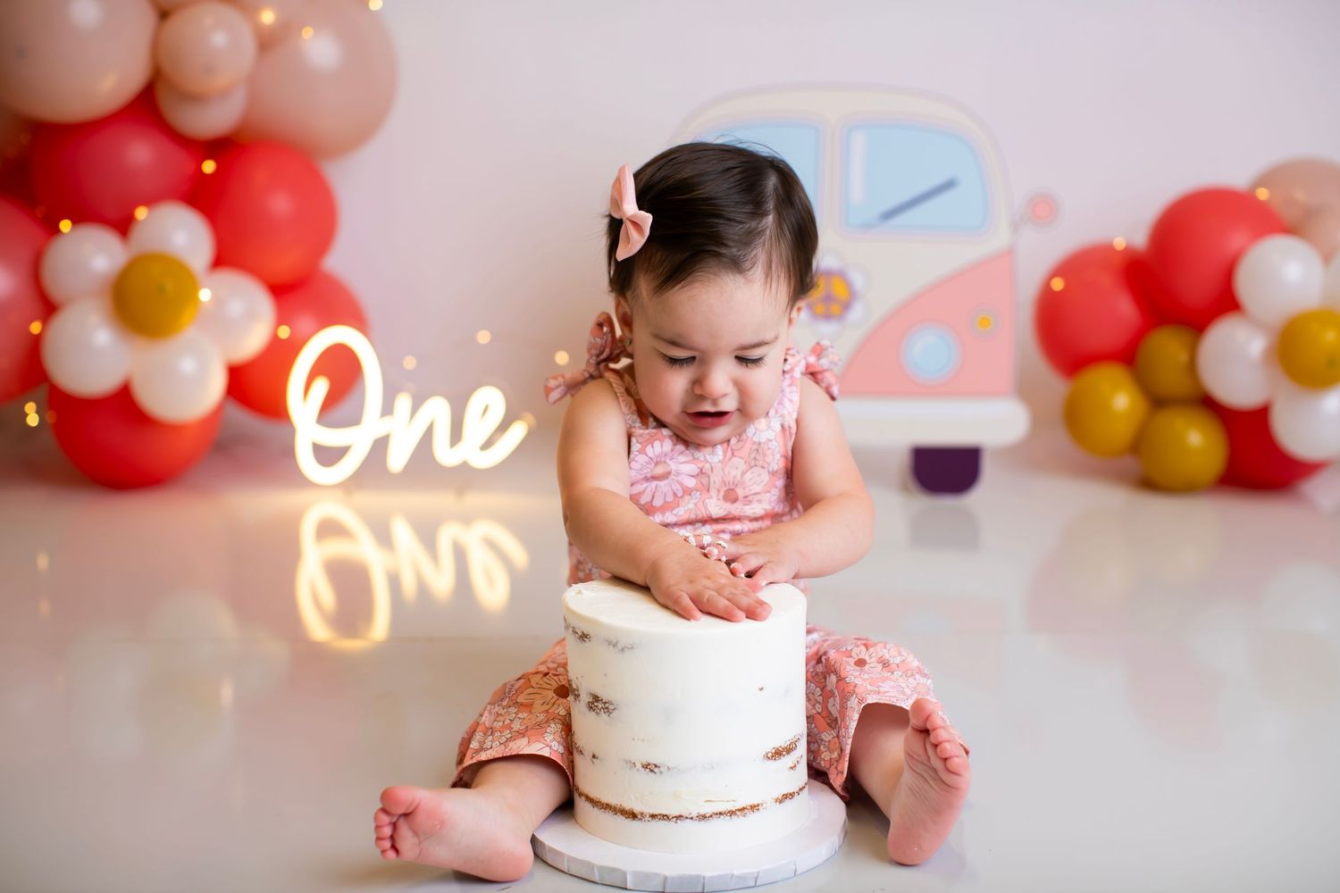 Baby in pink romper looking down at white birthday cake with balloon decorations.