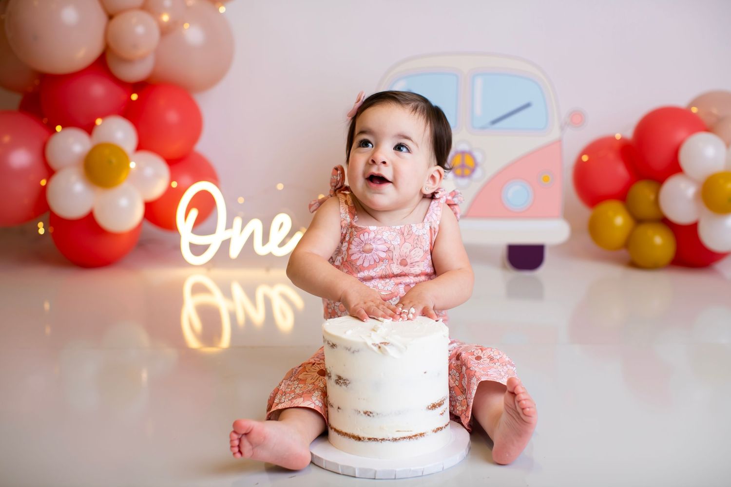 Baby looking up with excitement next to white birthday cake against balloon backdrop.