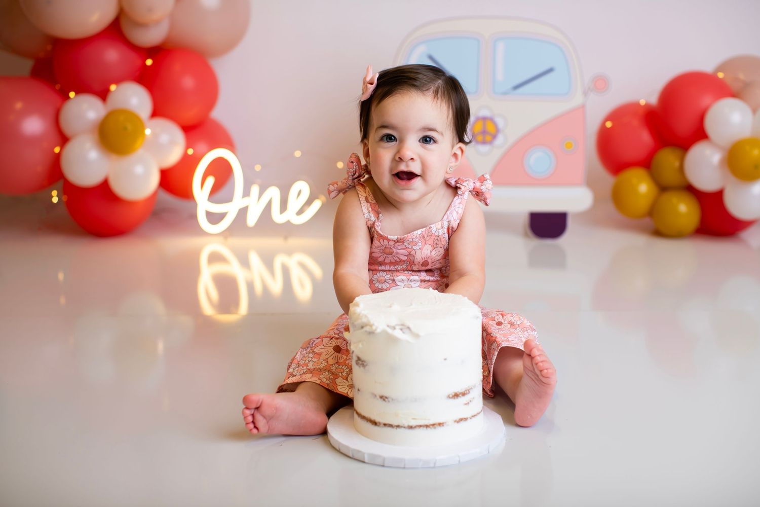 Happy baby sitting next to white birthday cake with balloon arch and neon sign in background.