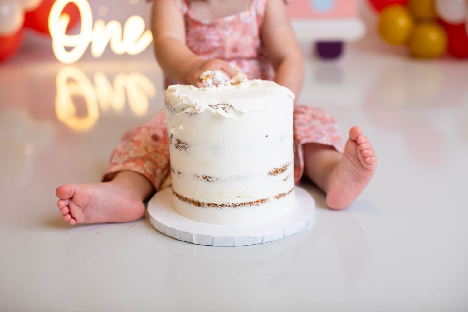 Tiny feet next to a white buttercream birthday cake with neon 'One' sign in the background.