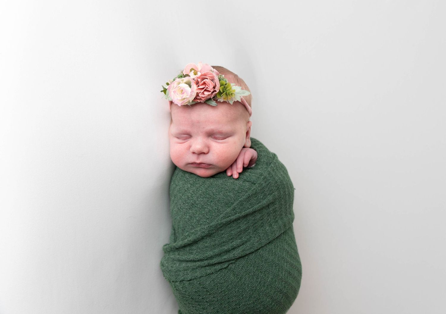 Newborn baby wrapped in green fabric wearing a delicate floral crown against white background.