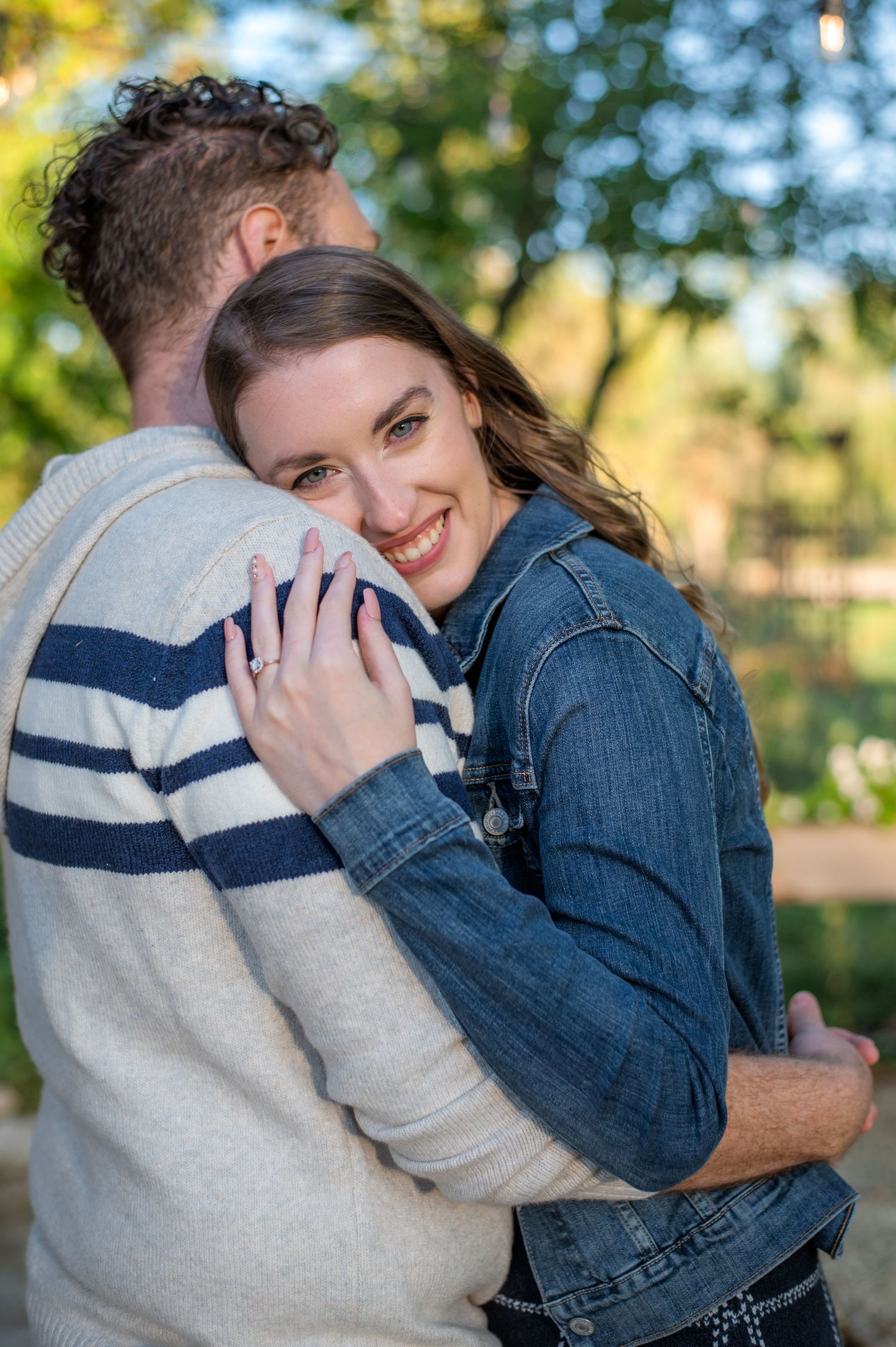 Couple embracing outdoors wearing coordinating sweater and denim jacket with natural backdrop.