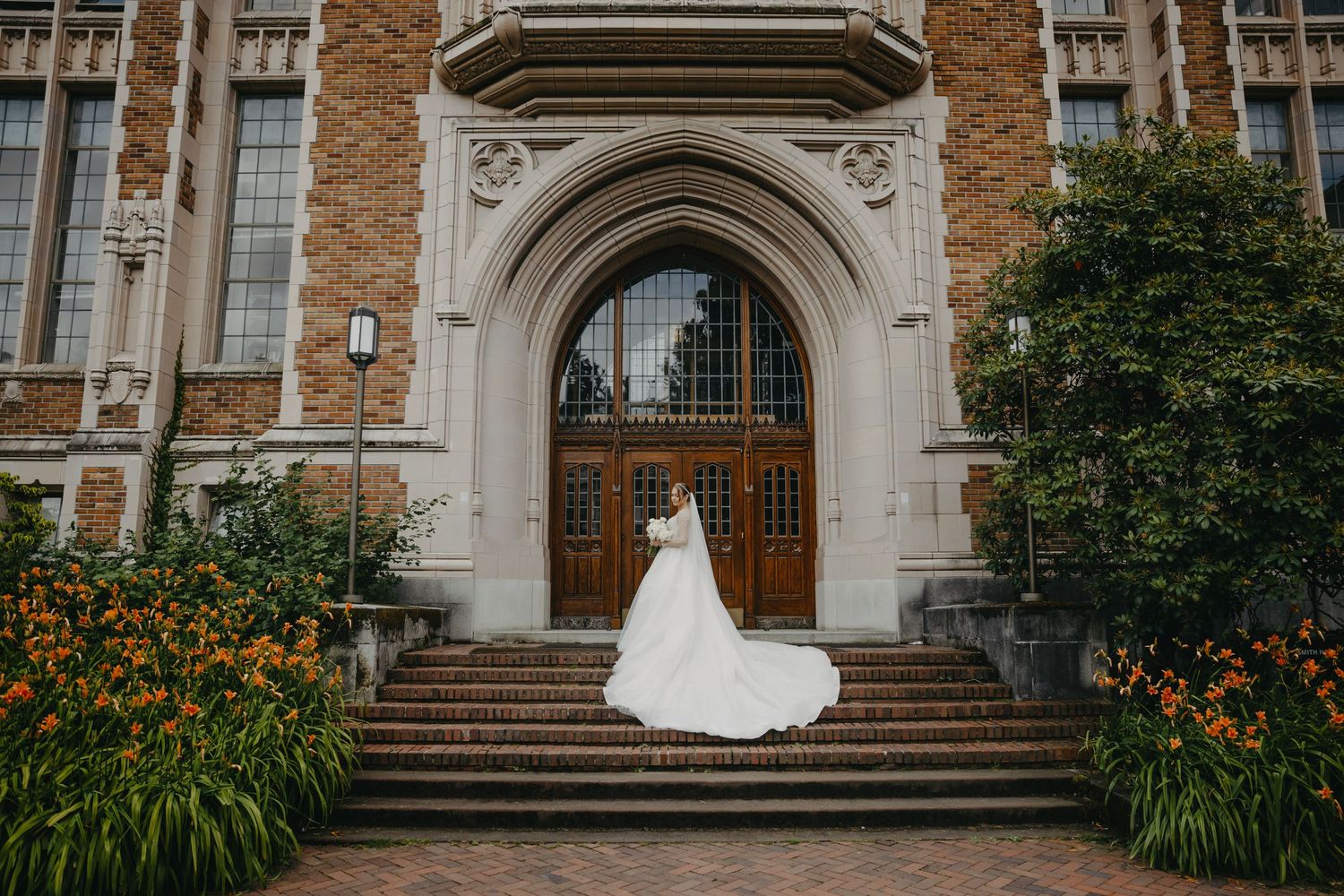 Bride poses on stone steps of historic church building with orange day lilies in foreground.