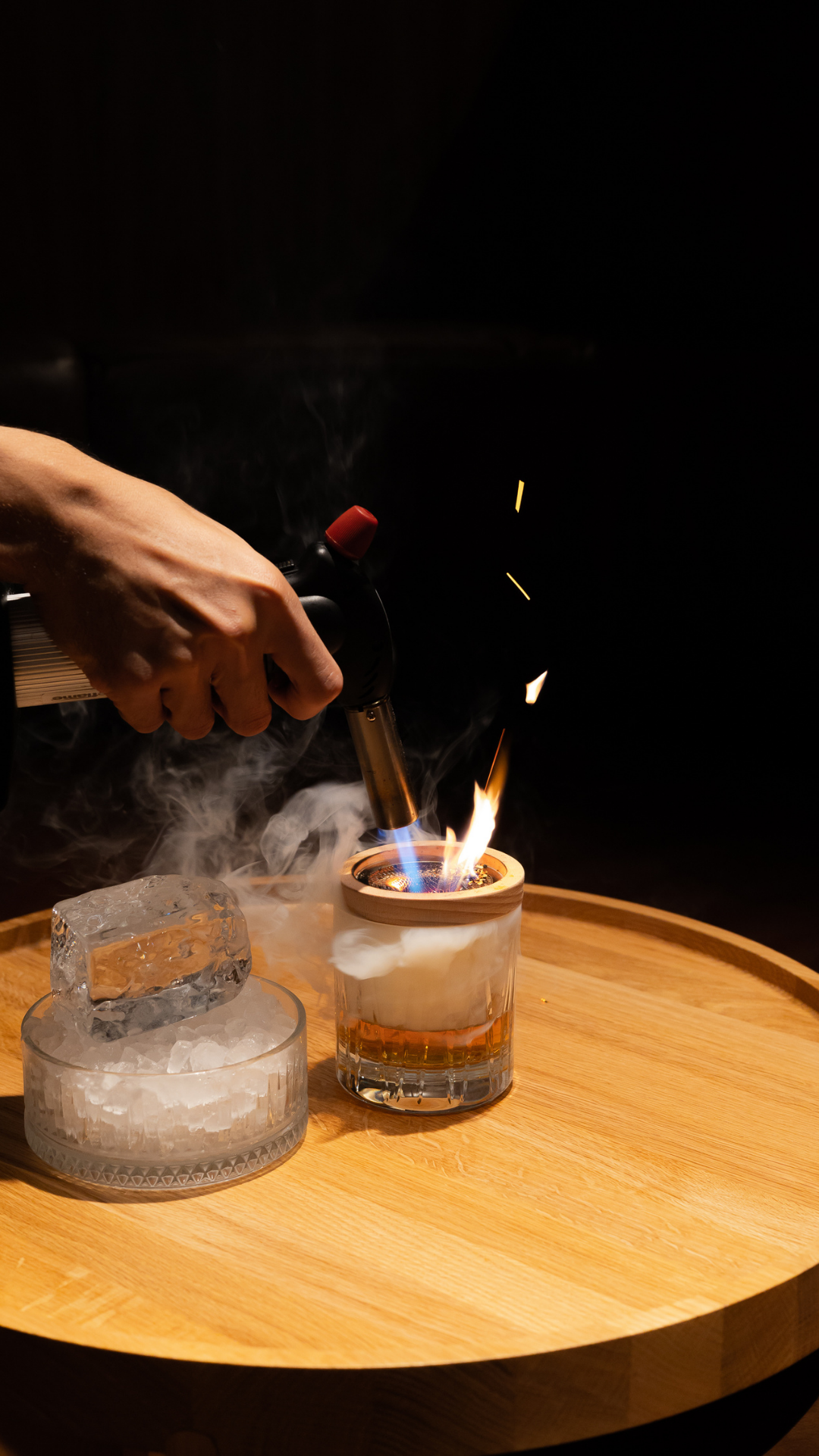 A dramatic cocktail presentation shows flames and smoke being created on a wooden circular tray.