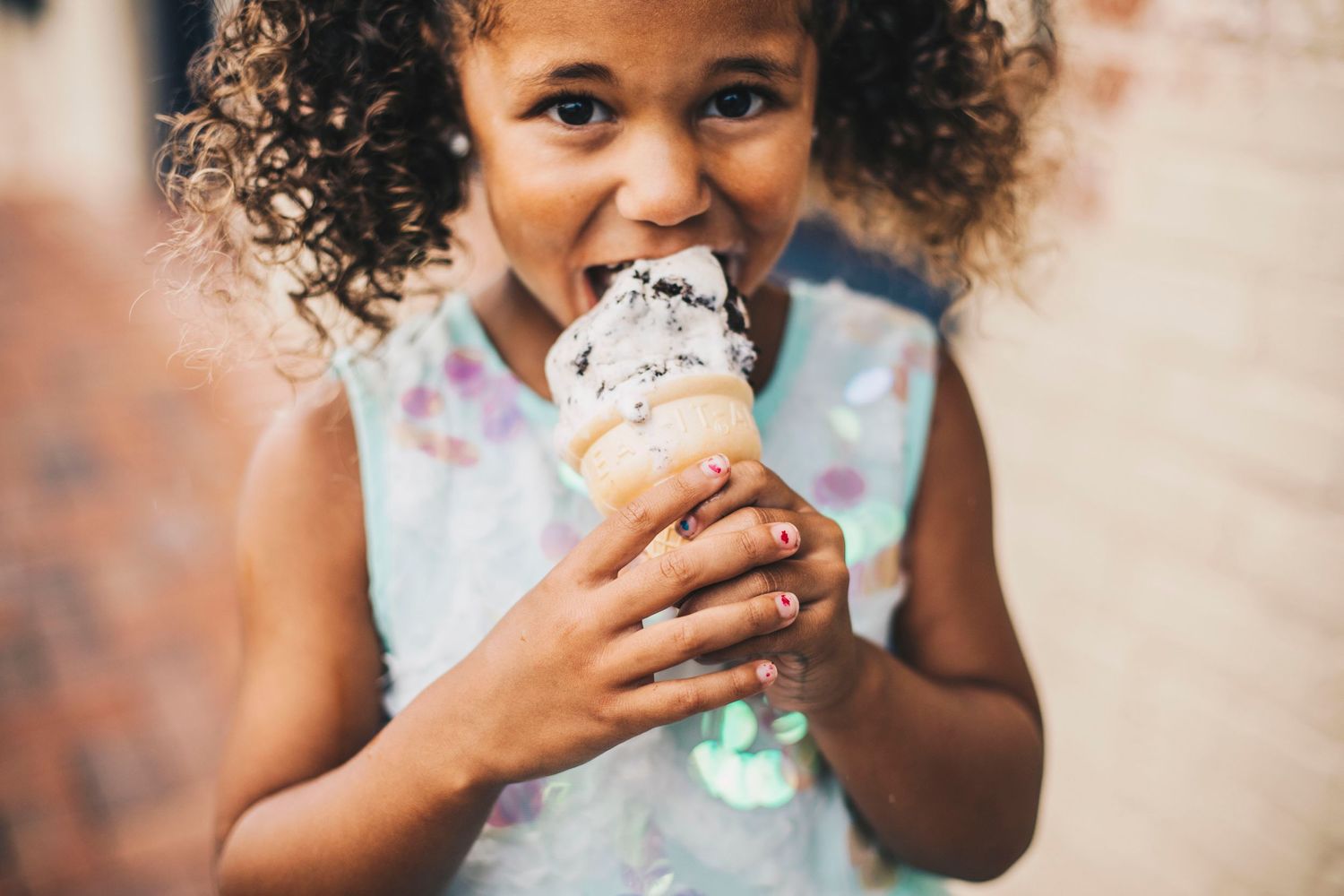 Young girl with curly hair eating a chocolate chip ice cream cone outdoors near a brick wall.