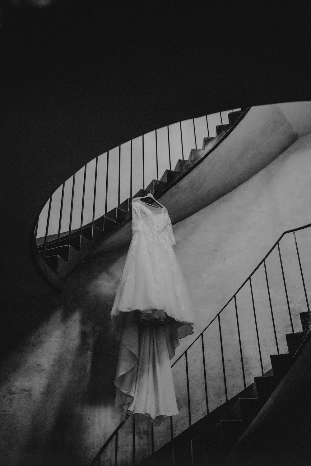 Dramatic black and white photo of wedding dress hanging on spiral staircase with natural light.