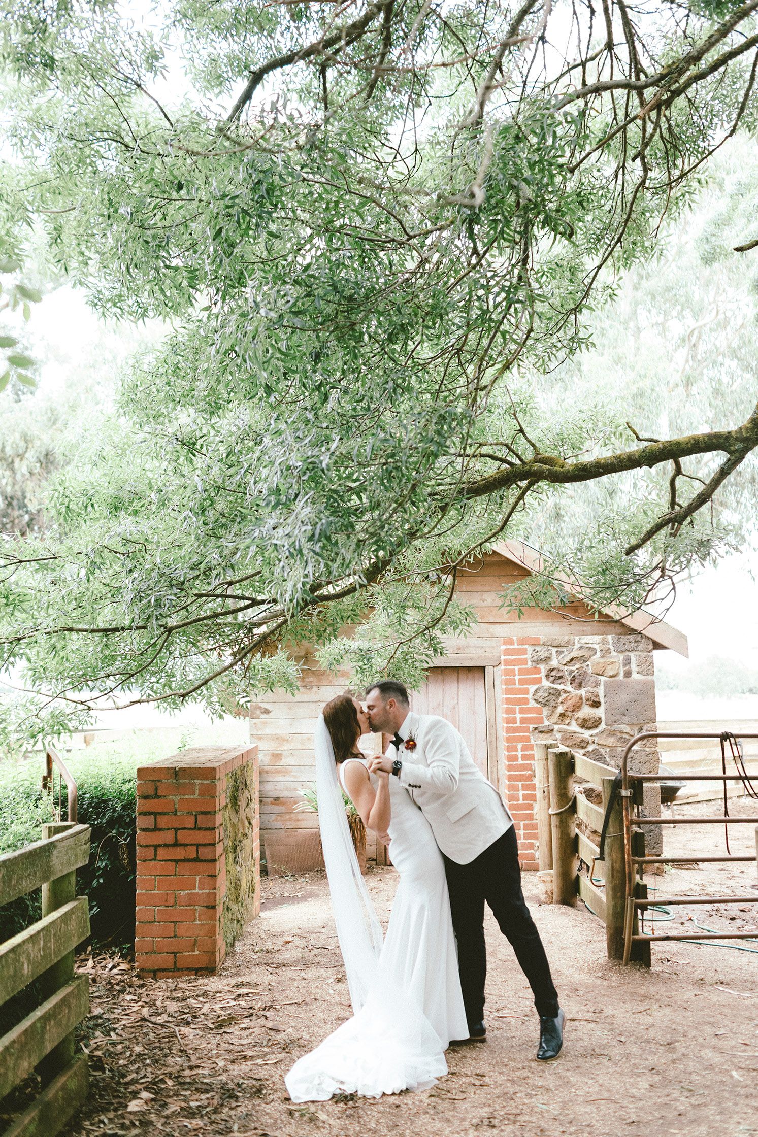 A romantic wedding moment under overhanging trees near a rustic brick and stone building.