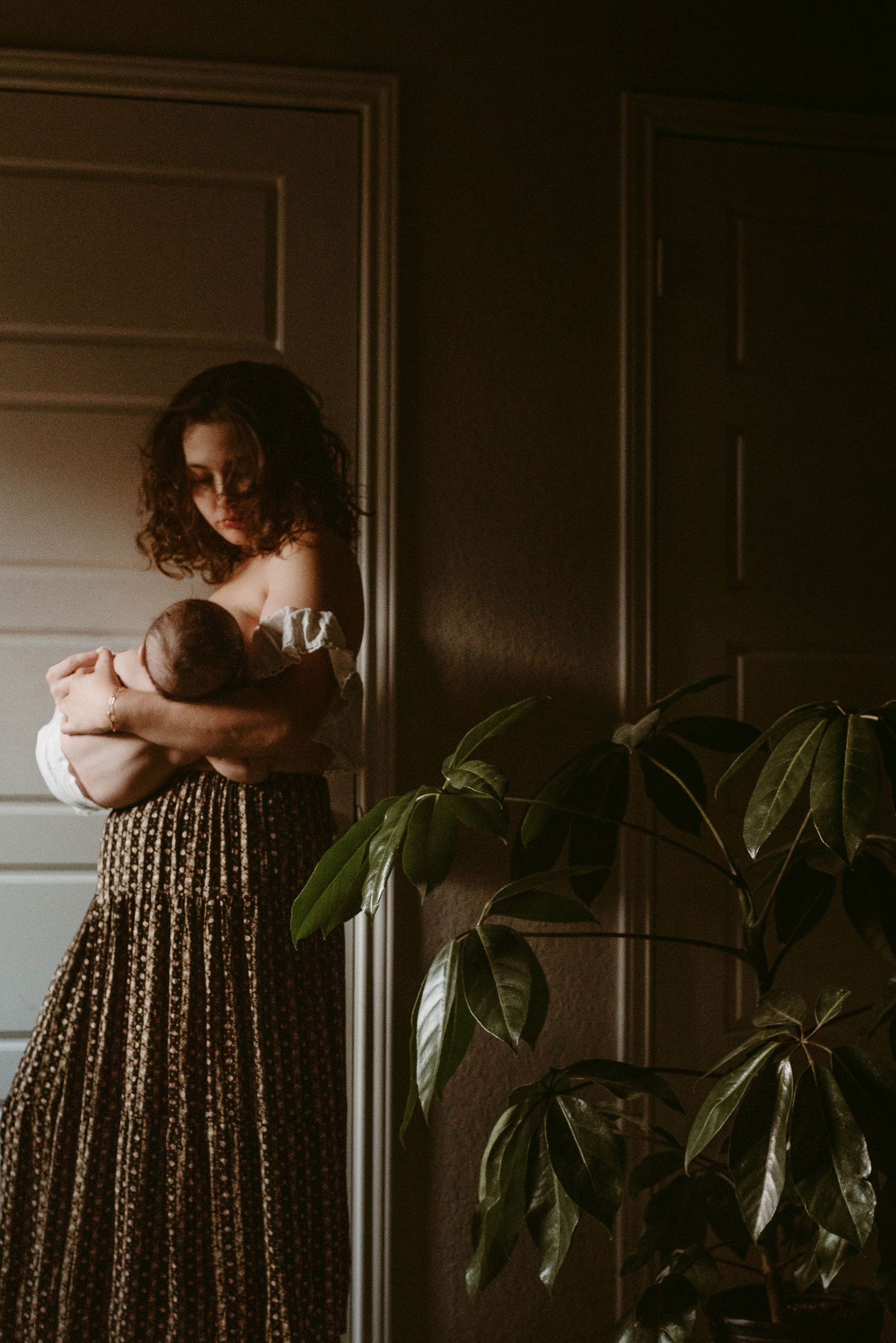 Moody indoor portrait with woman in patterned dress holding baby next to houseplant in dark setting.