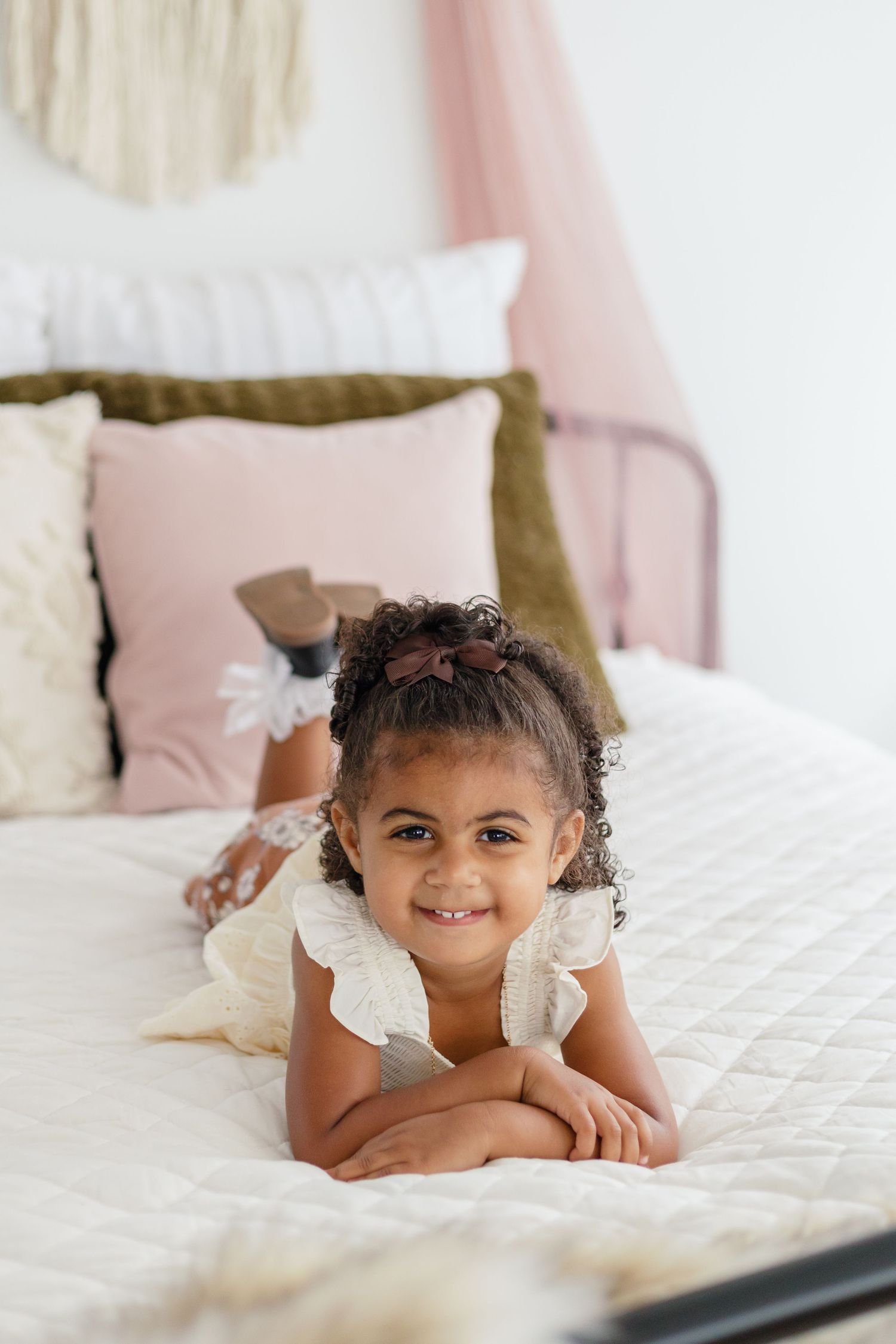 Child poses on white bed with pink and brown accent pillows and wall decor.